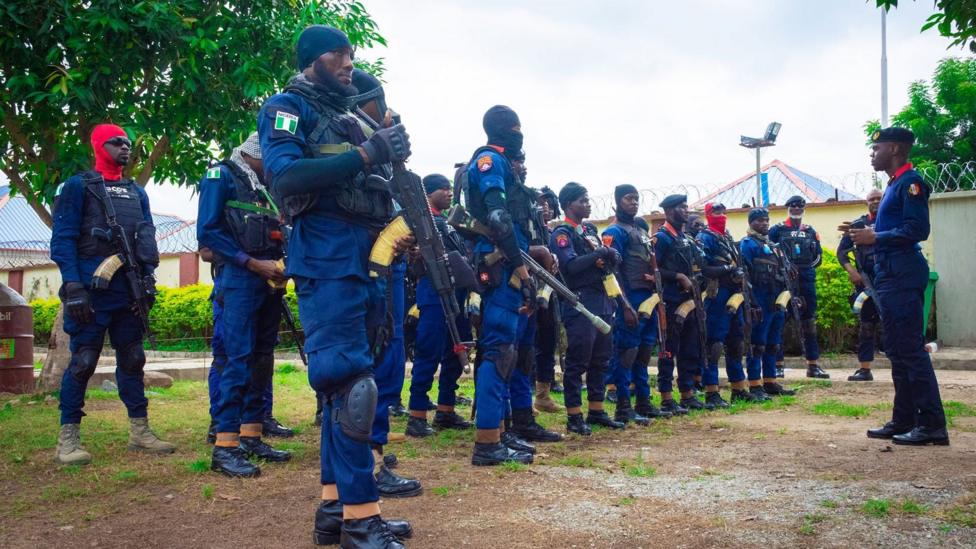 Two lines of armed men in blue uniform being addressed by a man wearing a beret
