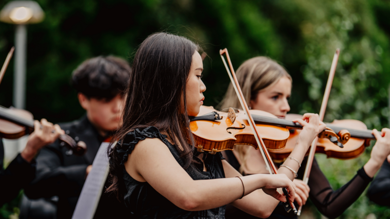 A number of people playing violins at Culture Night Belfast in 2025. There is a woman at the front with long brown hair and a dark blouse, playing a violin.