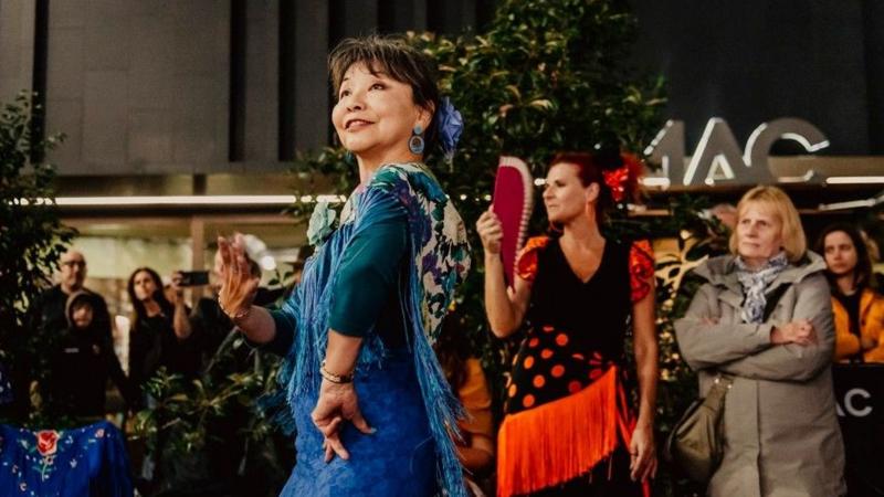A woman in a blue dress with dark hair dancing at Culture Night Belfast in 2025. There is a woman in a red and black dress to her right. There is a MAC sign in the background.