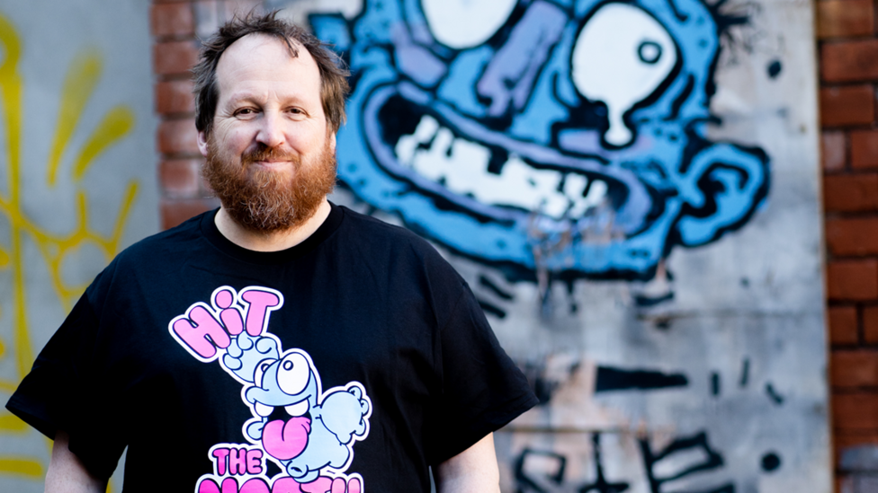 Adam Turkington, a man with brown hair and a beard. He is wearing a black t-shirt and standing in front of a wall with cartoon art on it.
