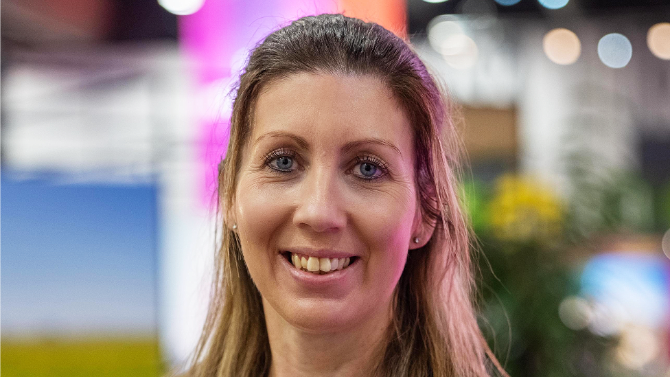 Head shot of Amanda Lloyd, a white woman in her 30s or 40s. She has long, partly bleached blonde hair with the front section tied back from her forehead. She has light blue eyes, wears clear stone stud earrings and is smiling directly at the camera. The background is interior, colourful but blurred.