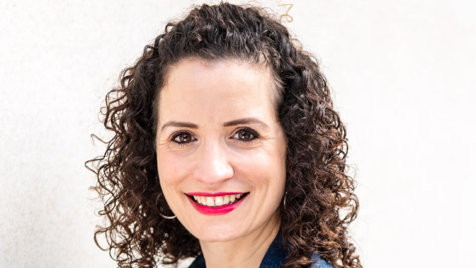 Emma Short is a white woman in her 40s. The photos shows her head only. She has dark brown eyes and dark, very curly neck-length hair which is pulled back off her forehead. The edge of a dark blue shirt can be seen. She is wearing make-up including bright pink-red lipstick and has silver hoop earrings. She is smiling directly at the camera with her head at a slight angle from her body.