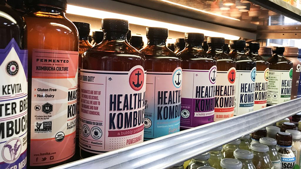 Dark brown bottles of kombucha on a shop shelf. Most of the products are called Health-Ade Kombucha and there are different coloured labets depending on the flavour, eg pomegranate and blood orange, with a ship's anchor logo above the name. Two bottles of Kevita Master Brew Kombucha can also be seen to the left of the photo.