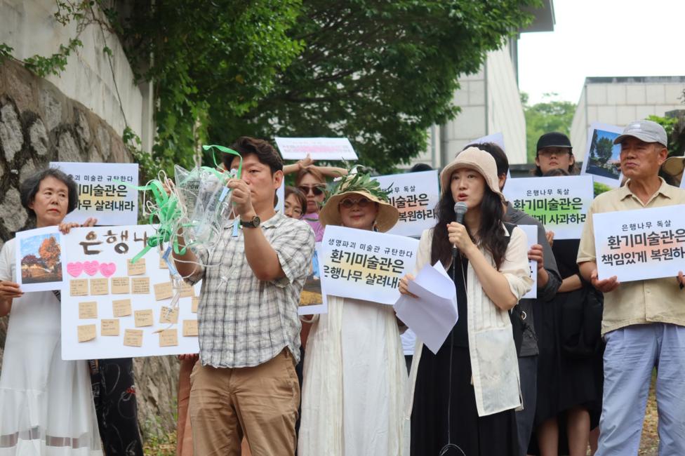 A group of people stand on a quiet, tree-lined lane holding handwritten signs and posters with Korean text. One person speaks into a microphone, while others display messages on placards. The scene is framed by stone and brick walls, and a large tree with a mixture of green and brown leaves behind the people.