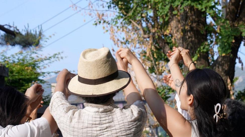Close-up view of several people standing outdoors beneath tall trees, raising their joined hands overhead in a circle. One person in the centre wears a light-colored straw hat, while others extend their arms toward the middle of the group.