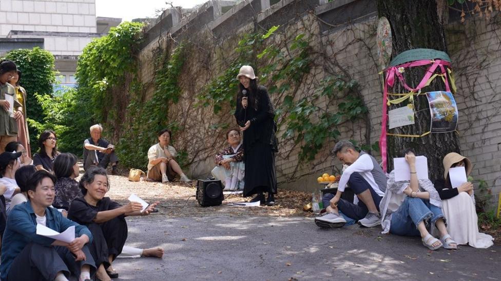 A group of about a dozen people sit and stand along a vine-covered brick wall. In the centre, a person dressed in black speaks into a microphone. To the right, a large tree is decorated with colorful ribbons, photographs, and papers.