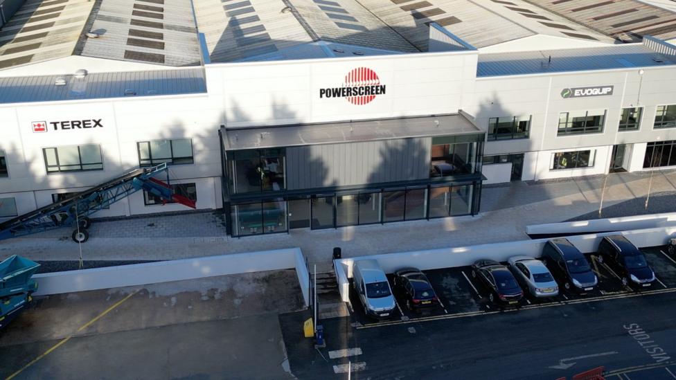 A large grey building which says powerscreen on it. There are cars parked outside it 