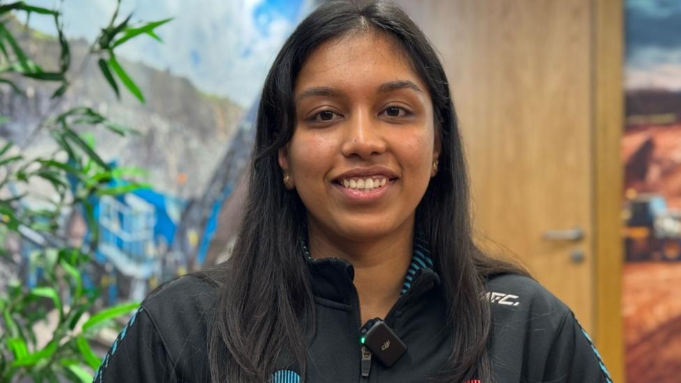 A young woman with long black hair and a sports jacket on smiles at the camera. 