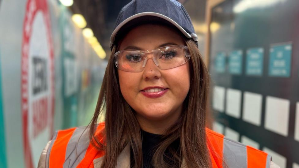 A brown haired woman with protective glasses , berry coloured lipstick and a high vis jacket looks at the camera with a slight smile. 