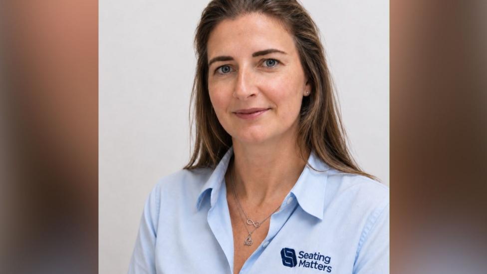 A woman with long brown hair, and two silver necklaces in a blue shirt stares at the camera. 