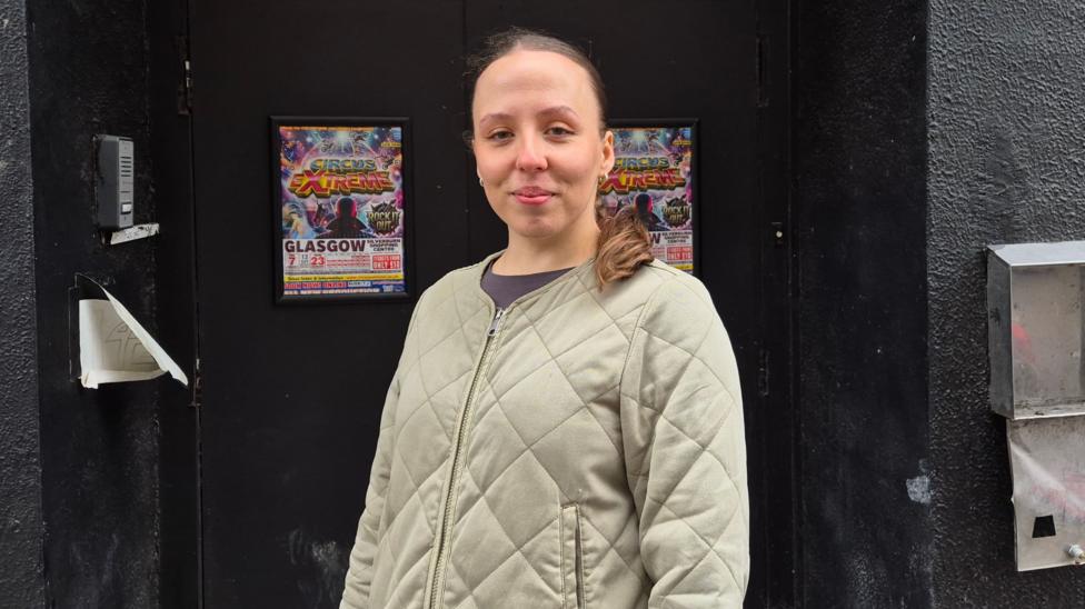 Nella McNicol standing in the doorway of a club. She has brunette hair, is wearing a grey or green jacket and is smiling into the camera.