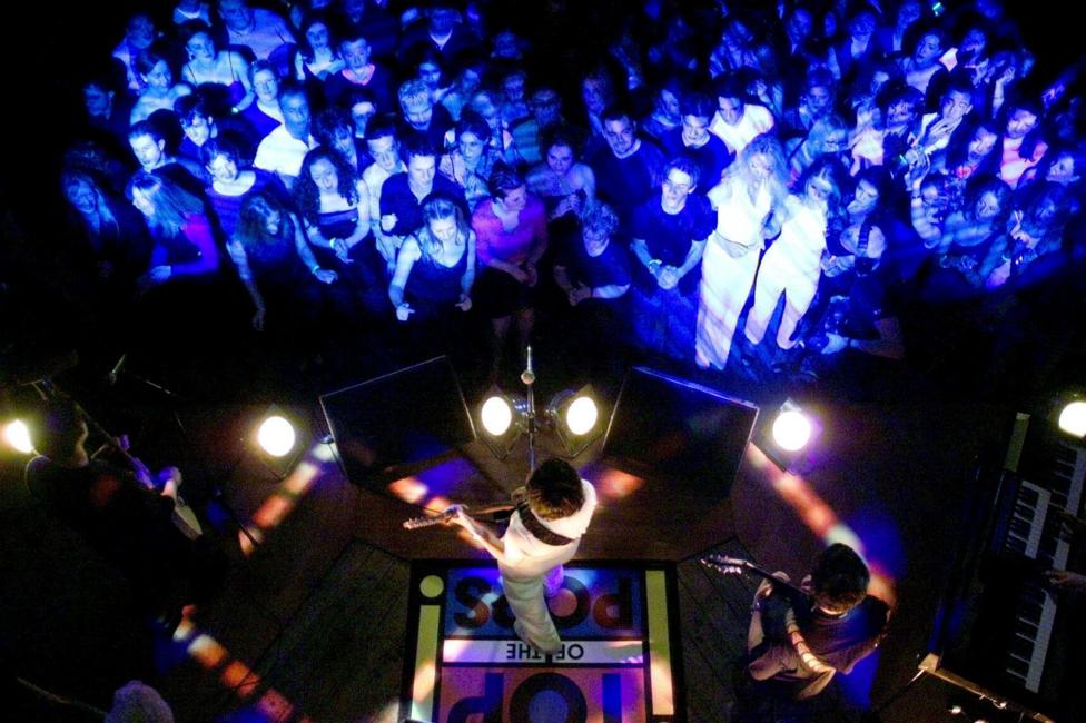 A band perform onstage in a club, watched by a crowd of people. On the stage floor is a banner saying Top of the Pops.