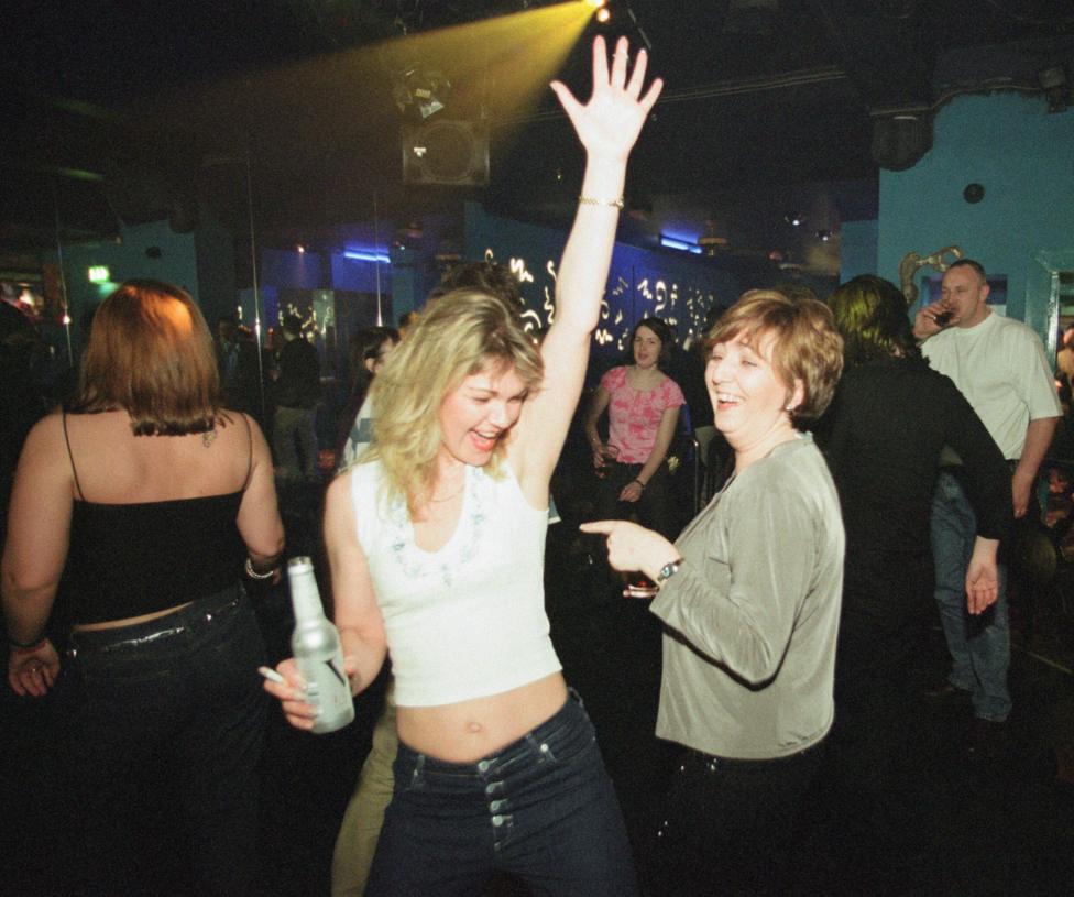 A woman dancing in a nightclub, laughing as she waves her hand in the air. Another woman next to her is laughing as she does so.