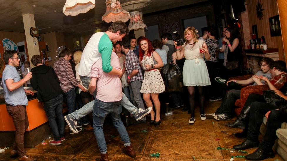 A small club, with people sitting drinking, standing by a bar or milling on the dancefloor. One man has jokingly jumped on another.