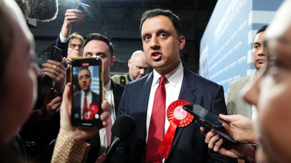 A person in a dark navy suit, white shirt, and bright red tie stands at the centre of a tightly packed media scrum. A large red rosette is pinned to the suit jacket, suggesting a political or election-related event. Several journalists and photographers surround the central figure, extending microphones and smartphones towards them. One smartphone in the foreground is actively recording or photographing the scene, with the central figure visible on the phone's screen.