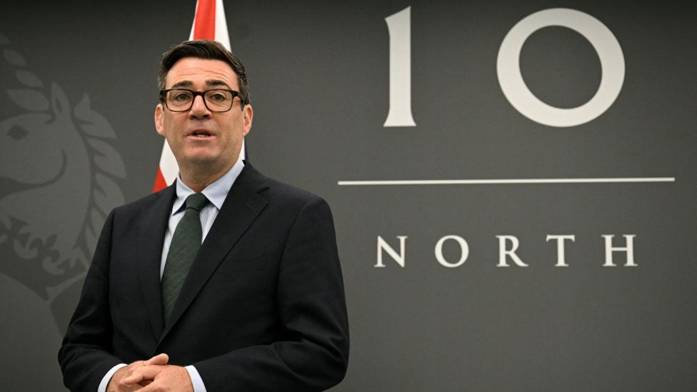 A person in a dark suit and glasses stands in front of a large “10 North” backdrop, with part of a red-and-white flag visible behind them.