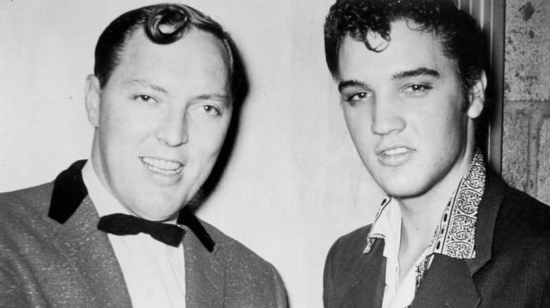 Black and white image of singers Bill Haley and Elvis Presley, which was taken in the 50s. They are both wearing suit jackets. Elvis is wearing an open-collared patterned shirt while Haley is wearing a white shirt with a dark bow tie.