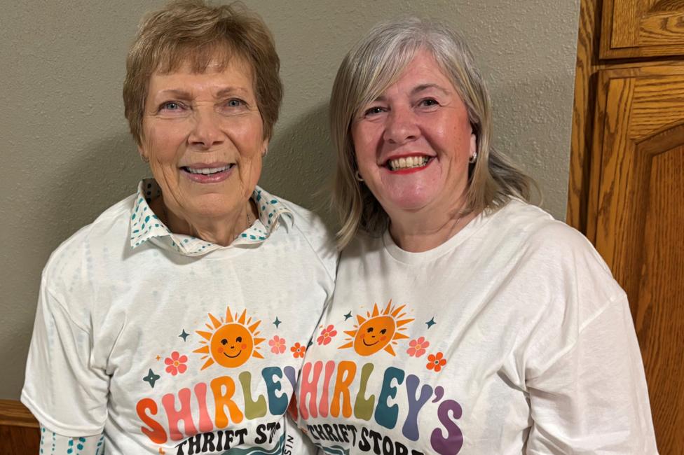 The two Shirleys are wearing the same T-shirt. It is white with a smiley son and colourful writing on it that says Shirley's thrift store