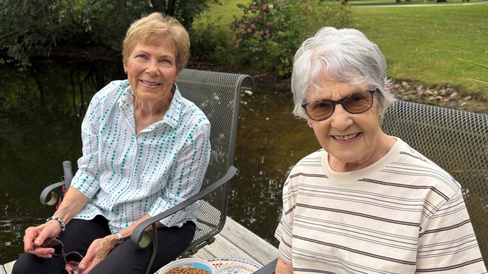 Shirley Yaegar and Kathleen Wilson are sitting in a garden and are both smiling at the camera. Shirely has short, brownish hair and is wearing black trousers and a white and blue patterened shirt. She is wearing a bracelet and holding sunglasses. Kathleen has short, grey hair and is wearing sunglasses. She si wearing a beige and brown striped T-shirt.