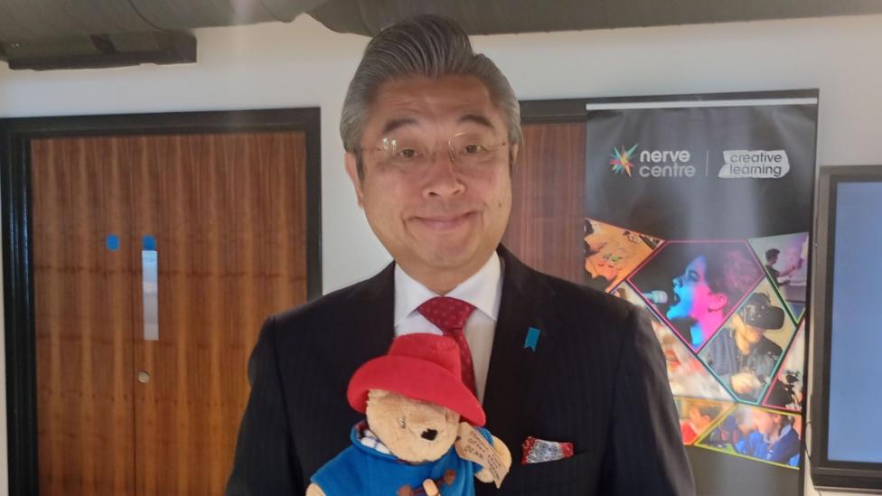 A man in a black suit, white shirt and red tie is holding a teddy bear with a blue coat and red hat.