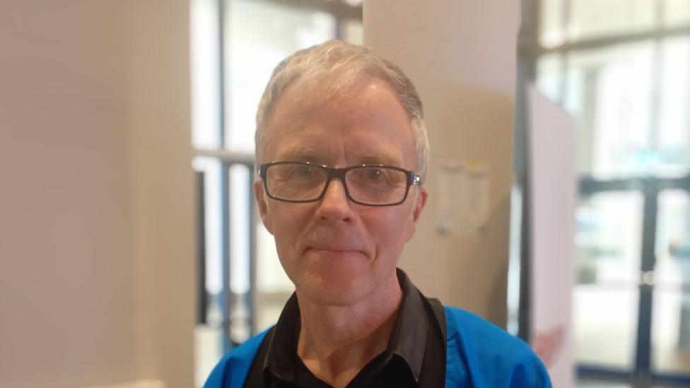 A man with black glasses and grey hair. He is wearing a black shirt and blue coat.