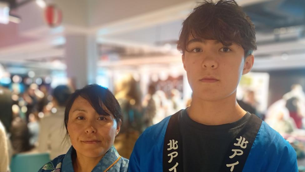 A woman stands next to a boy. She is wearing light blue clothes. He is wearing dark blue clothes with a black lapel. There is white writing on the lapel.