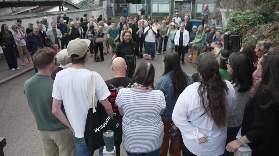 A large group of men and women standing in a circle, at a train station. A man stands in the centre appearing to conduct or lead a song.