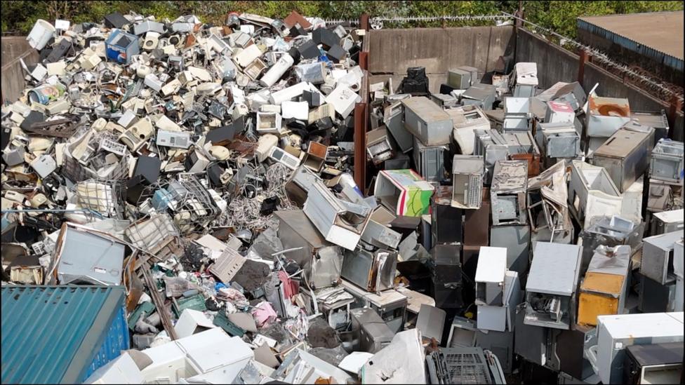 A pile of appliances such as fridges and freezers sit in a fenced off area.