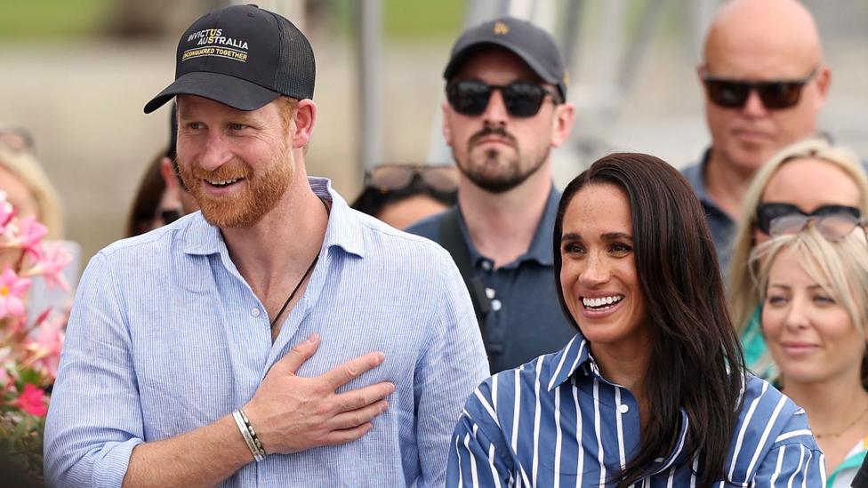 Harry and Meghan seen walking together in Sydney harbour in April. He is wearing a blue shirt and a cap, she is wearing a striped blue shirt.