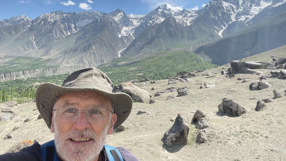 Sean Drysdale who has a grey beard and steel rimmed glasses and a floppy hat takes a selfie with a spectacular range of snow-capped mountains behind him in the Chundu Valley in Pakistan