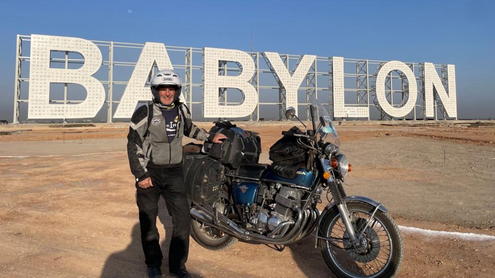 Sean Drysdale is wearing biker gear and a helmet and standing in front of his motorbike in front of a large white sign that says Babylon on it