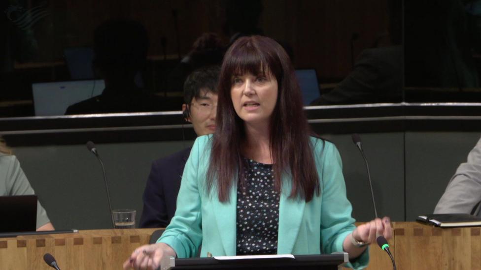 Helen Jenner at First Minister's Questions in the Senedd chamber. She is standing behind a lectern and has long dark hair with a fringe. She is wearing a light turquoise blazer, a dark top with a white speckled or polka-dot pattern. Papers or notes are placed on the lectern. Three other Senedd members are partially visible. 