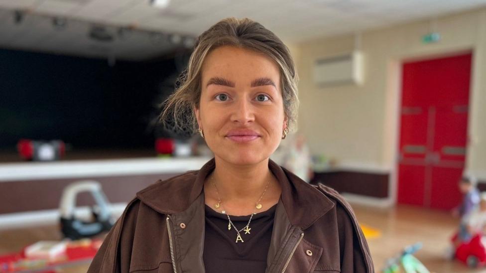 Amelia is pictured at a toddler group in a community centre. She has brown hair which is tied back from her face in a ponytail. She is wearing a black t shirt and a brown jacket and gold coloured jewellery.