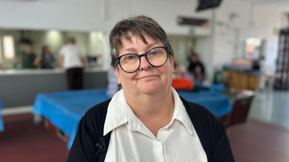 Penny is pictured in the canteen of a community centre. She has dark hair and is wearing black rimmed glasses. She is dressed in a white shirt and black cardigan.