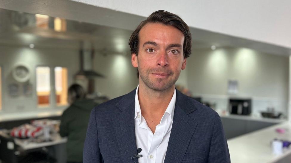 Andrew is pictured in the kitchen of a community centre. He is wearing a dark blue suit. He has dark brown hair and brown eyes.