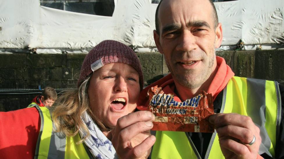A woman stands next to a man with her mouth open in astonishment as he holds an old, crumpled Marathon-branded chocolate wrapper in his hands.