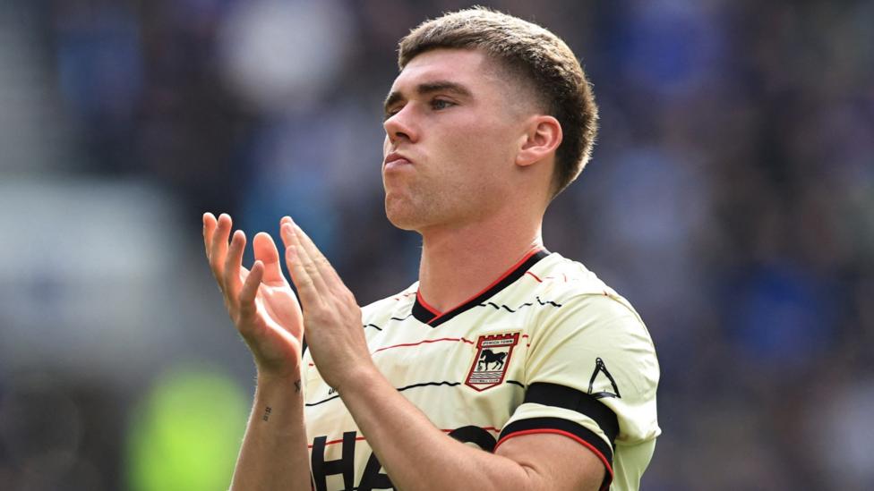 Ipswich Town player Leif Davis stands and applauds. He is wearing the club's cream coloured away kit with black and red horizontal pinstripes and a black armband on his left bicep.