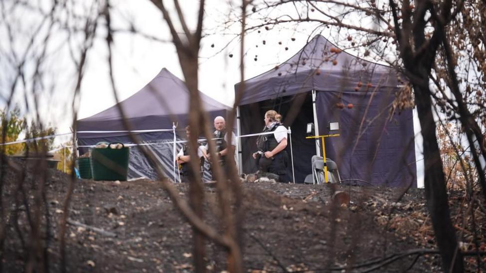 Two temporary tents and several people inside a taped-off area on a hillside with dark ground and sparse vegetation in the foreground.