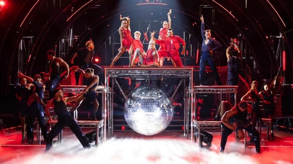 Five red-clad dancers on top of a metal contraption, surrounded by other backing dancers in black.