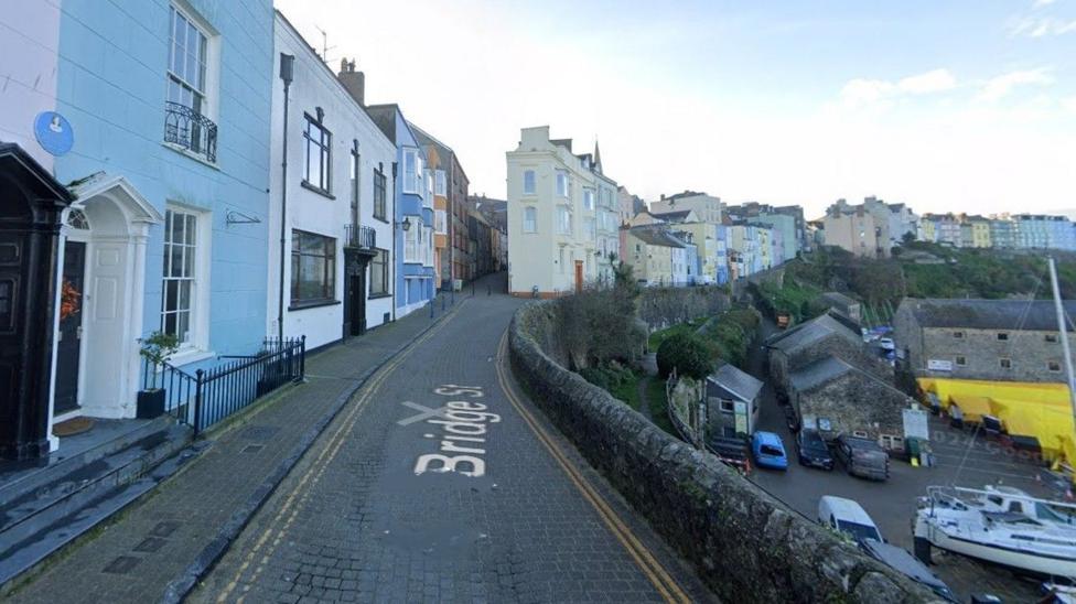 Google street view of Bridge Street in Tenby. There is a row of different coloured homes on a narrow cobbled street