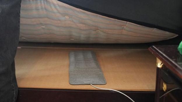 A slim rectangular grey pad with a white wire coming out the end is laid on a wooded board and you can see a mattress being held up above it.