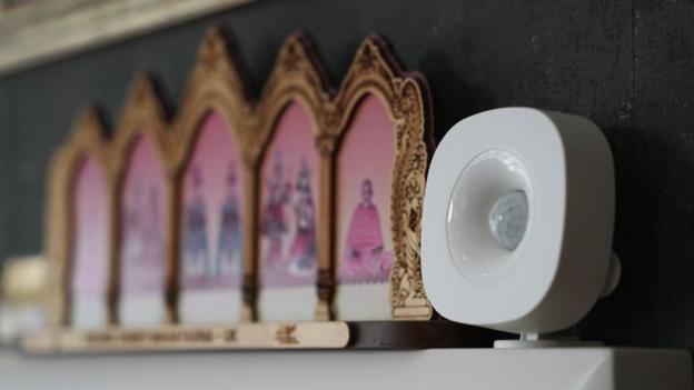 A small white round device with an almost globe shape in the middle sits on a shelf with an ornament showing hindu gods in the background which are blurred.