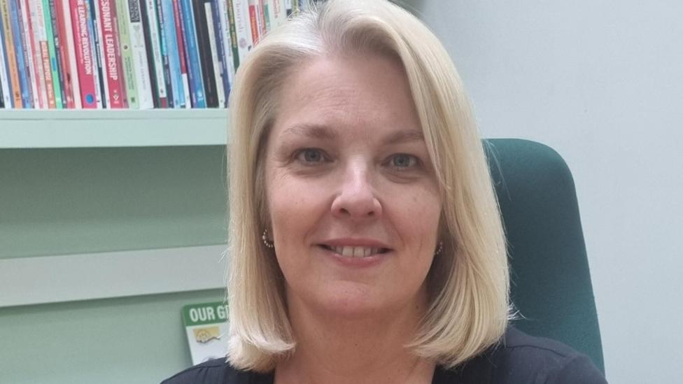 Headteacher Amanda Hulme smiling at the camera in a school office. She has medium-length blonde hais and is wearing hooped earrings. She is sitting on a green chair and behind her is a bookshelf filled with books.