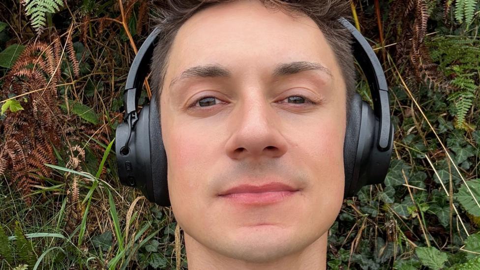 James Downs is lying on grass and wearing headphones. He has short brown hair that is spiky on top.