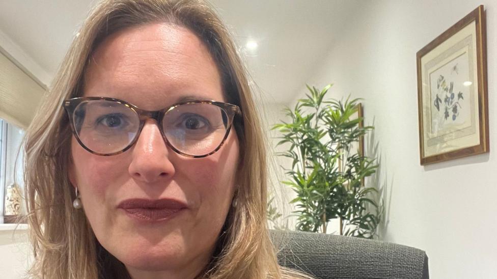 Lucy Theo has blonde shoulder-length hair and glasses. A plant and framed pictures of flowers can be seen behind her,