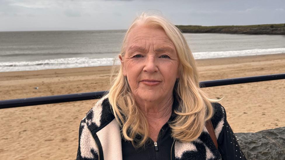 Susan Harvey has long blonde hair and is wearing a black and white fleece. The beach and sea can be seen behind her.