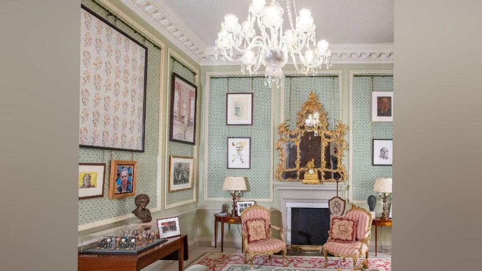 A room decorated in a mix of Georgian and Victorian style. The wallpaper is light green. The chairs are pink striped. The fireplace is grey. An ornate gold mirror hangs above the mantel. It features portraits of a number of well known political faces in NI's political history.