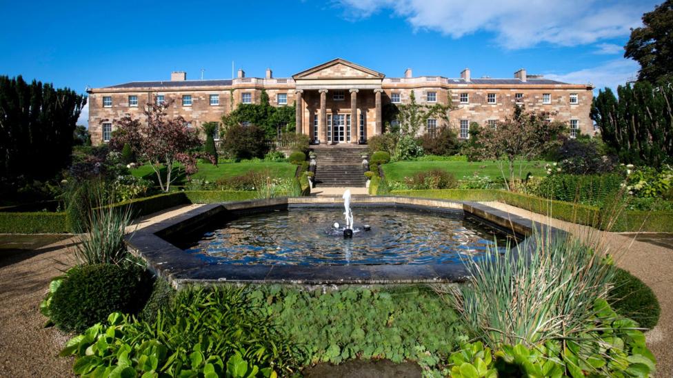 Northern Ireland's royal residence, Hillsborough Castle and Gardens. It is a long stone building with pillars at the front of it. Steps lead down to a landscaped garden with a water fountain at the front 