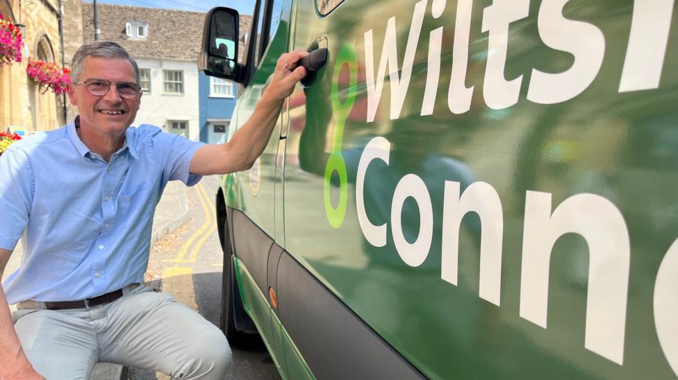 Martin Smith smiles at the camera. He is crouching next to the side of the Wiltshire Connect bus, his hand on the door handle and the Wiltshire Connect logo in view. He wears a blue shirt and light-coloured trousers.