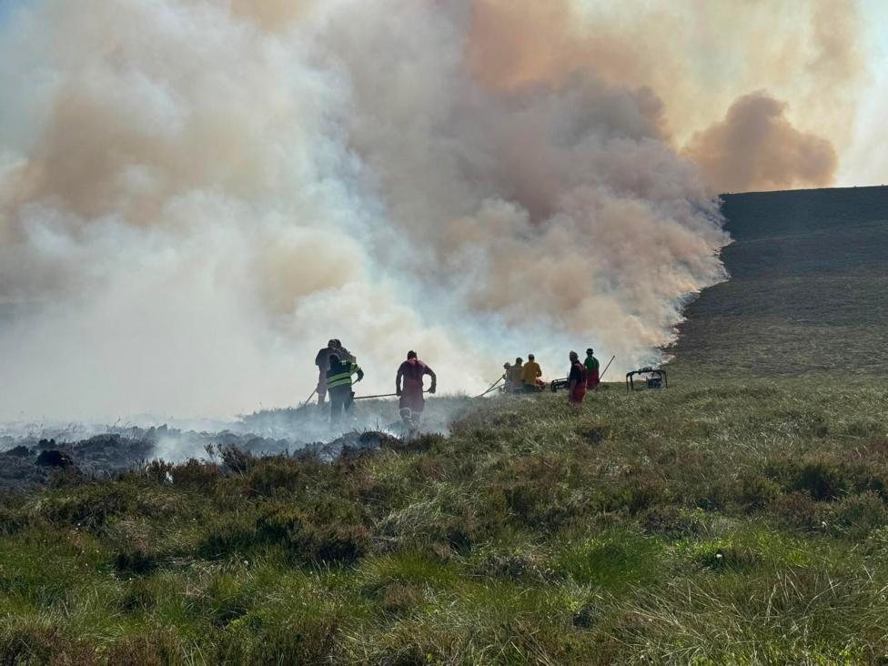 A group of people on a grassy moorland next to thick plumes of smoke.